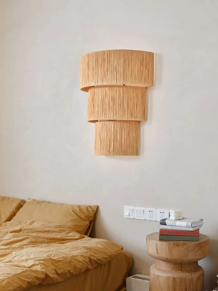 Natural raffia wall sconce light with tiered design, mounted above a bedside with mustard yellow bedding and a wooden stool.